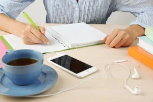 Persona escribiendo en una libreta con una taza de té, un smartphone y audífonos sobre el escritorio, representando el manejo del estrés laboral a través de la organización y pequeñas pausas.