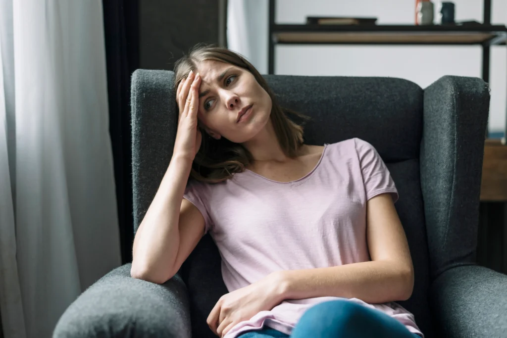 Mujer joven sentada en un sillón, con expresión preocupada y una mano en la frente, representando los síntomas de ansiedad.